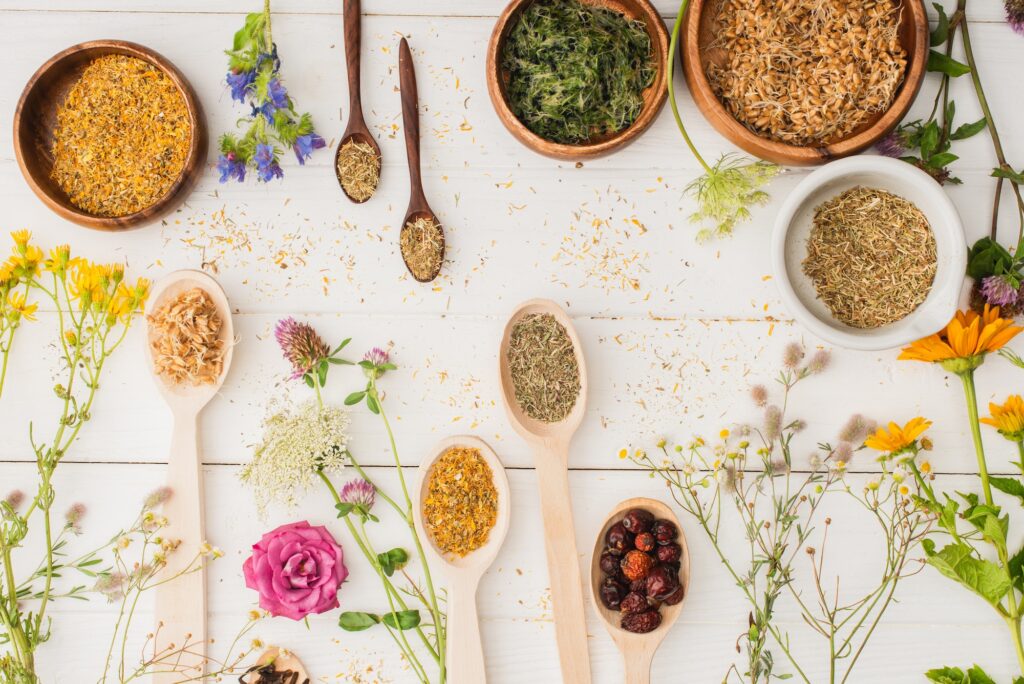 top view of herbs in spoons and bowls near flowers on white wooden background, naturopathy concept